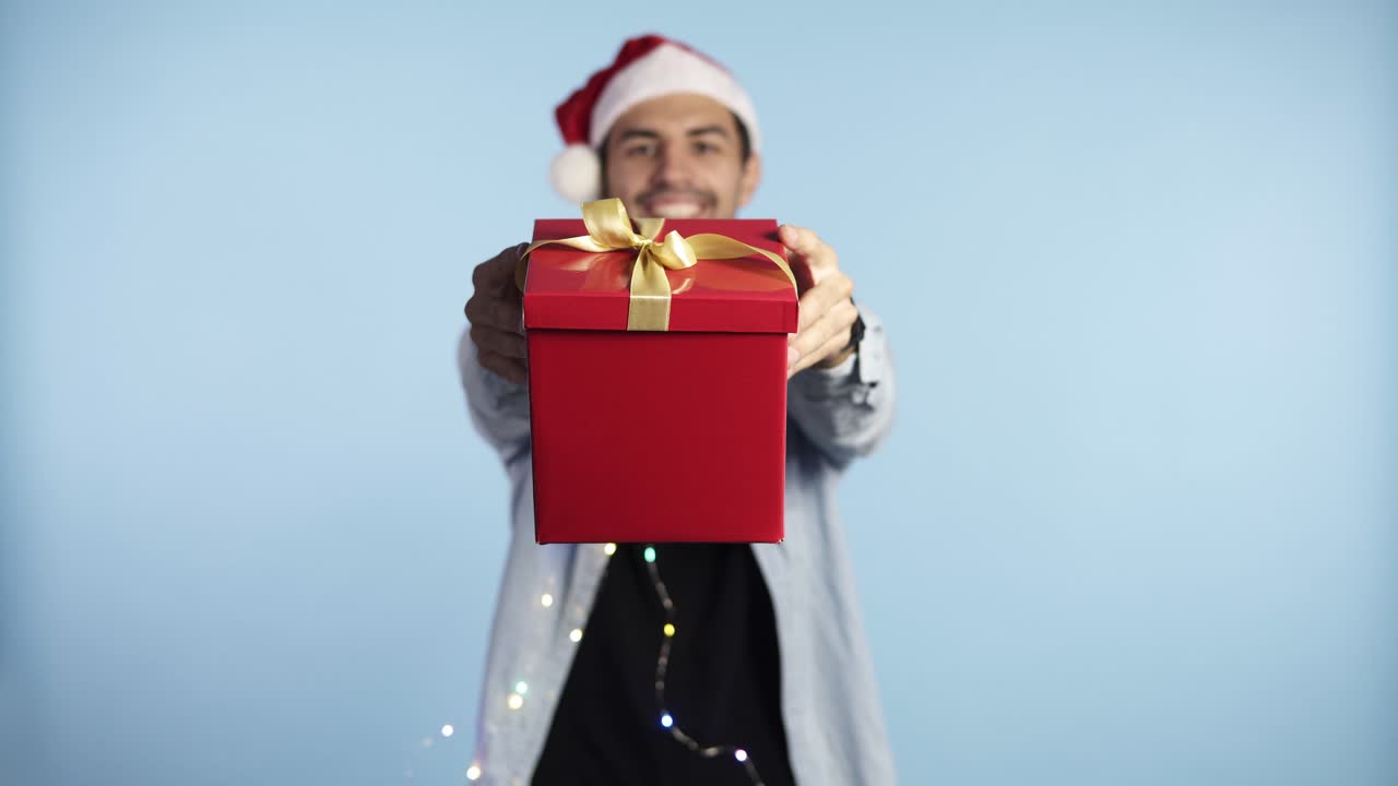 크리스마스 산타 모자와 화환을 목에 걸고 고립된 파란색 배경 위에 선물을 들고 손을 뻗은 채 행복한 얼굴로 서서 이빨을 보여주는 자신감 있는 미소를 짓고 있는 청년. 닫고, 선물 상자에 집중하세요