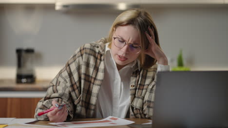 the woman works remotely from home sitting at a table with a laptop and a felt-tip pen marks the data on the graph. nervously talking on the video link