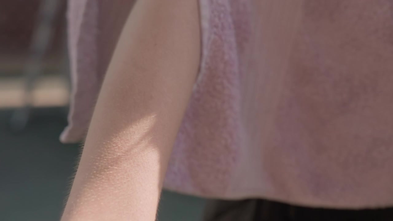 Close-up lengan dan tangan wanita saat mengeringkan diri dengan handuk merah muda, memperlihatkan kuku yang terawat dengan cat kuku berwarna terang. Pencahayaan lembut menonjolkan tekstur kulit dan kain dalam suasana luar ruangan.