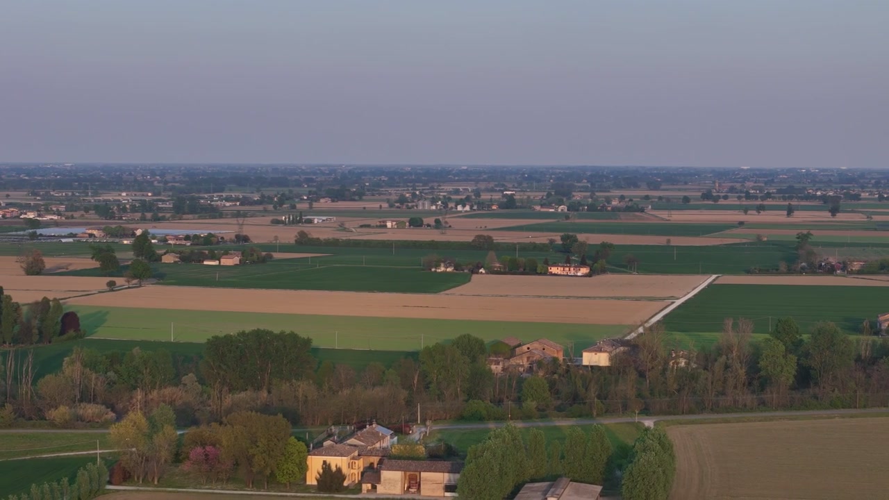 Ripresa aerea con drone che si allontana sopra i campi agricoli della Pianura Padana e gli appezzamenti alberati con insediamenti rurali sparsi, rivelando la struttura piatta del paesaggio della pianura coltivata.