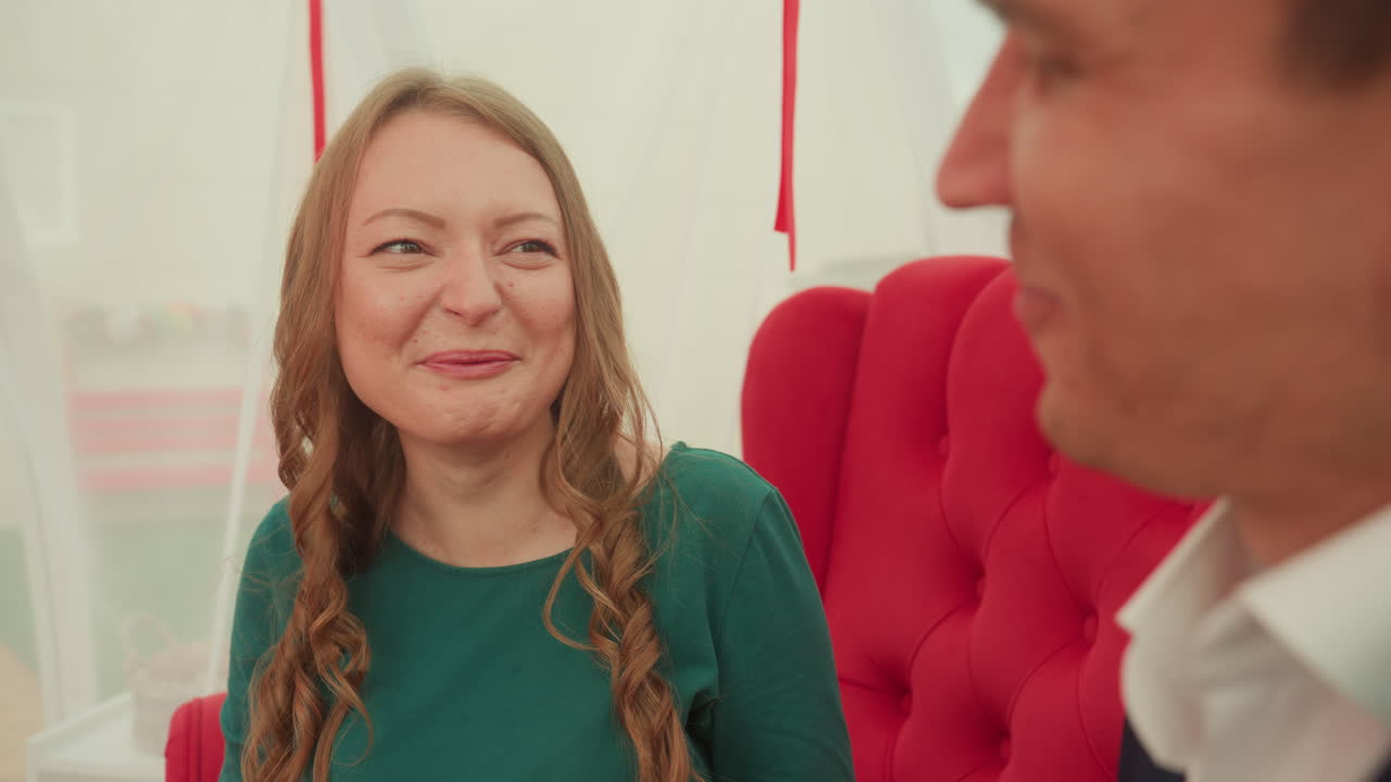 coppia bianca su divano rosso che assaggia. Un uomo in giacca e cravatta imbocca delicatamente una donna mentre lei sorride, contatto visivo intimo e morso giocoso, sfondo in velluto trapuntato, abito verde casual e giacca, luce naturale soffusa