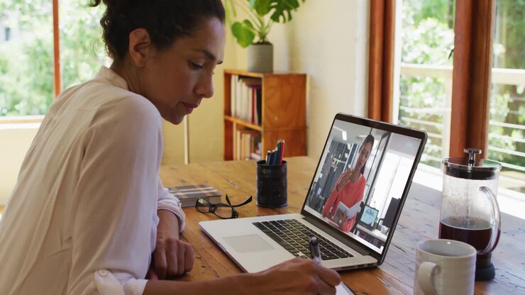 Afroamerikansk kvinde tager notater under et videoopkald på bærbar computer derhjemme.