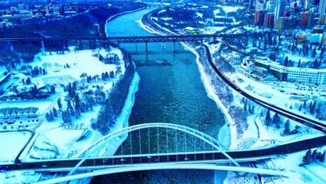 Vista-Aérea-De-Invierno-De-4.000-Aves-Sobre-El-Puente-Walter-Dale-Mientras-Los-Pedazos-De-Hielo-Fluyen-Por-El-Río-Saskatchewan-Del-Norte,-Con-El-Antiguo-Puente-Antiguo-De-Alto-Nivel-En-El-Horizonte,-Por-Victoria-Y-Kinsmen-Parks2-3