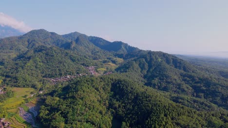 Aerial-view-of-tropical-rural-landscape-of-forest,-hills-and-village-against-blue-sky