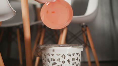 close up of person seated on modern chair using hand to drop fresh peeled carrot strips into patterned waste bin lined with plastic bag on wooden floor in cozy home kitchen setting