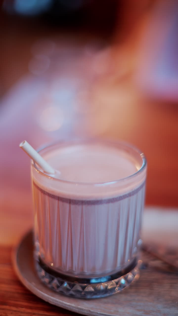 Close up of a hot chocolate in a glass with a straw on a table at a cafe. Vertical