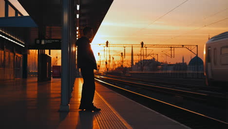 man waiting on a train platform at sunset with a train passing by