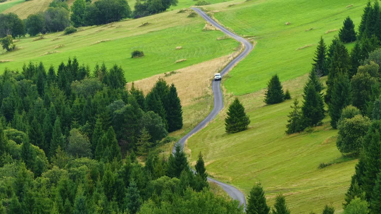 Aerial view of a vehicle traveling along a winding rural road surrounded by green fields and forest in Slovakia, showcasing peaceful countryside scenery