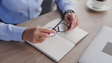 Female hands holding the glasses. Woman puts off the glasses and closes her paper notebook. Close up.
