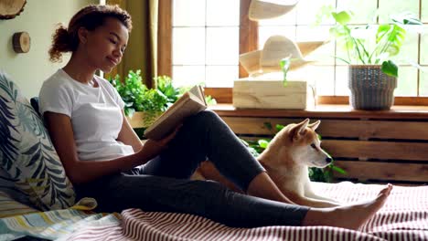 attractive girl is reading book and stroking beautiful dog lying on bed near its owner at home. modern interior with large window, green plants and wooden decor is visible.