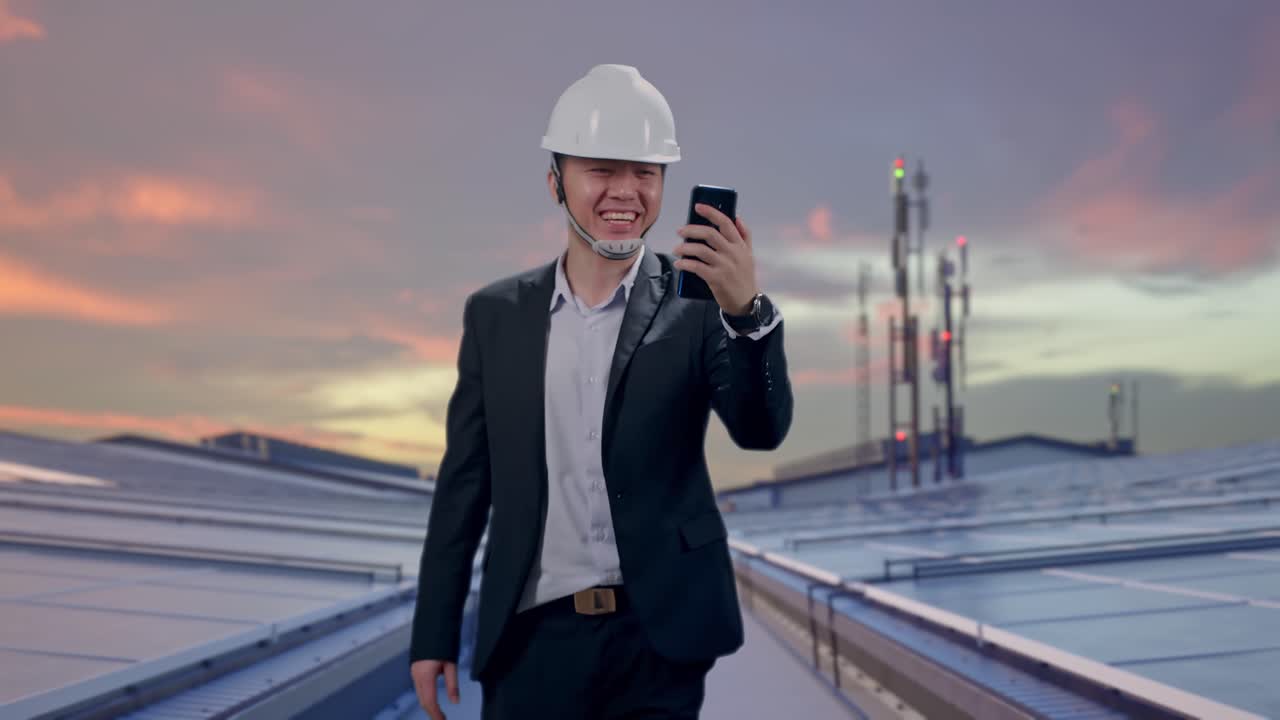 Vista frontal de empresario asiático usando un smartphone, en videollamada, mientras camina en un tejado industrial con paneles solares.