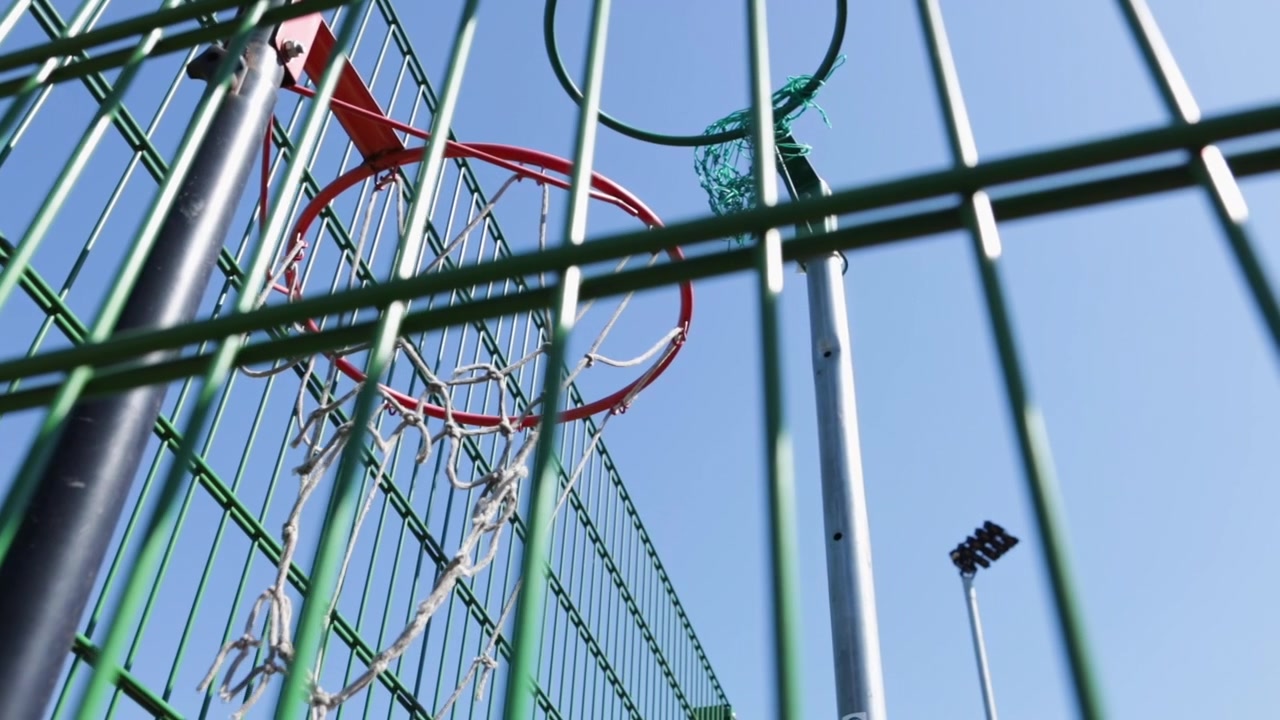 En rustik basketkorg med ett slitet nät står bakom ett grönt stålstaket på ett utomhussportområde. Låg vinkelbild.