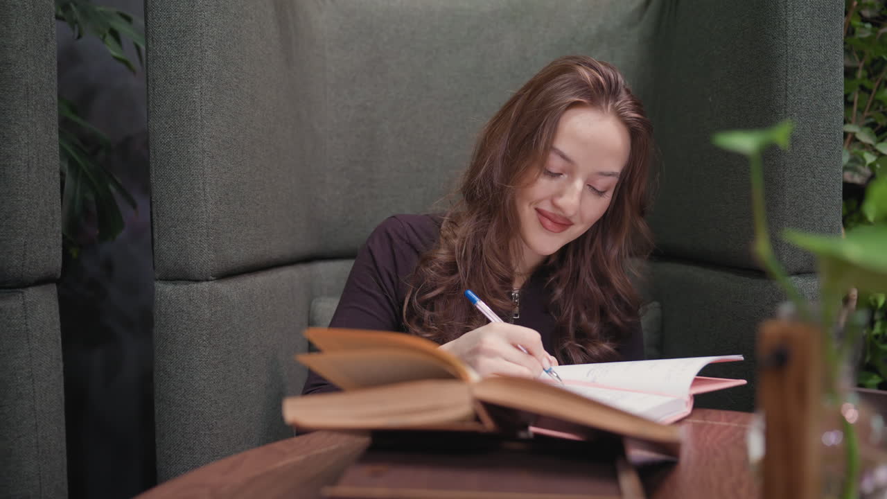 Lady with long wavy hair writes in pink notebook while smiling gently, seated in cozy booth with open book, decorative vase with green leaf, and relaxing indoor greenery in soft background light