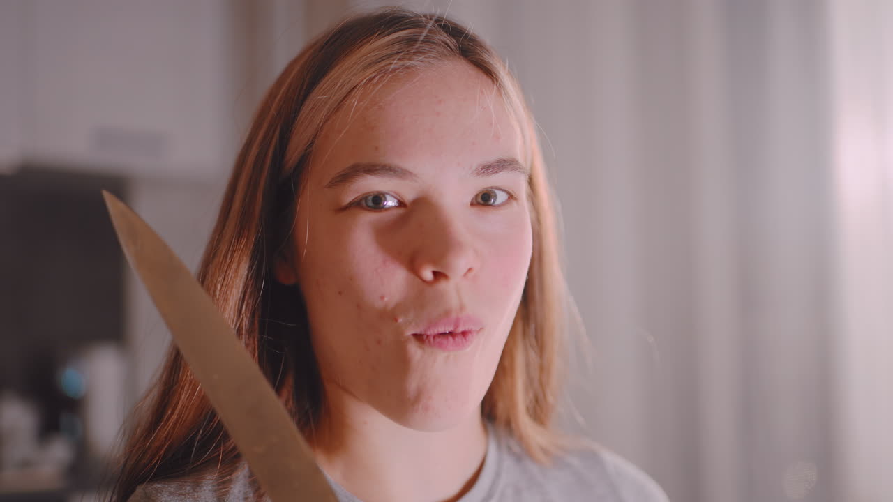 Smiling woman in kitchen holding knife after slicing cucumber, looking at camera with relaxed expression, illuminated by soft natural light while preparing fresh food in casual home environment