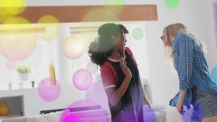 Animation of colourful light spots over two diverse teenage girls dancing at home