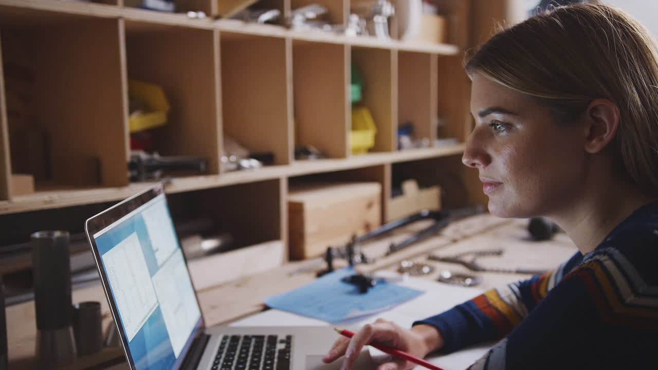 Close Up Of Female Engineer In Workshop Using Laptop And Making Notes On Plan For Bicycle
