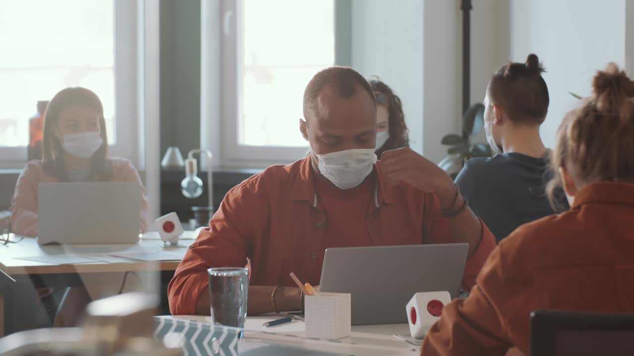 얼굴 마스크를 입은 아프리카계 미국인 남자가 오픈 스페이스 사무실에서 일하고 있습니다.