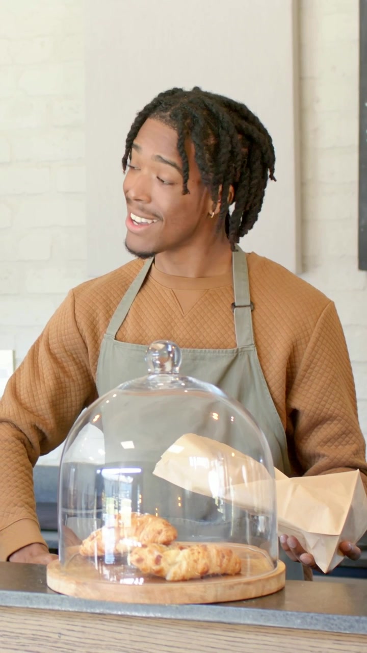 Vertical video: Holding bag, barista in apron packing croissant from cloche for customer at counter