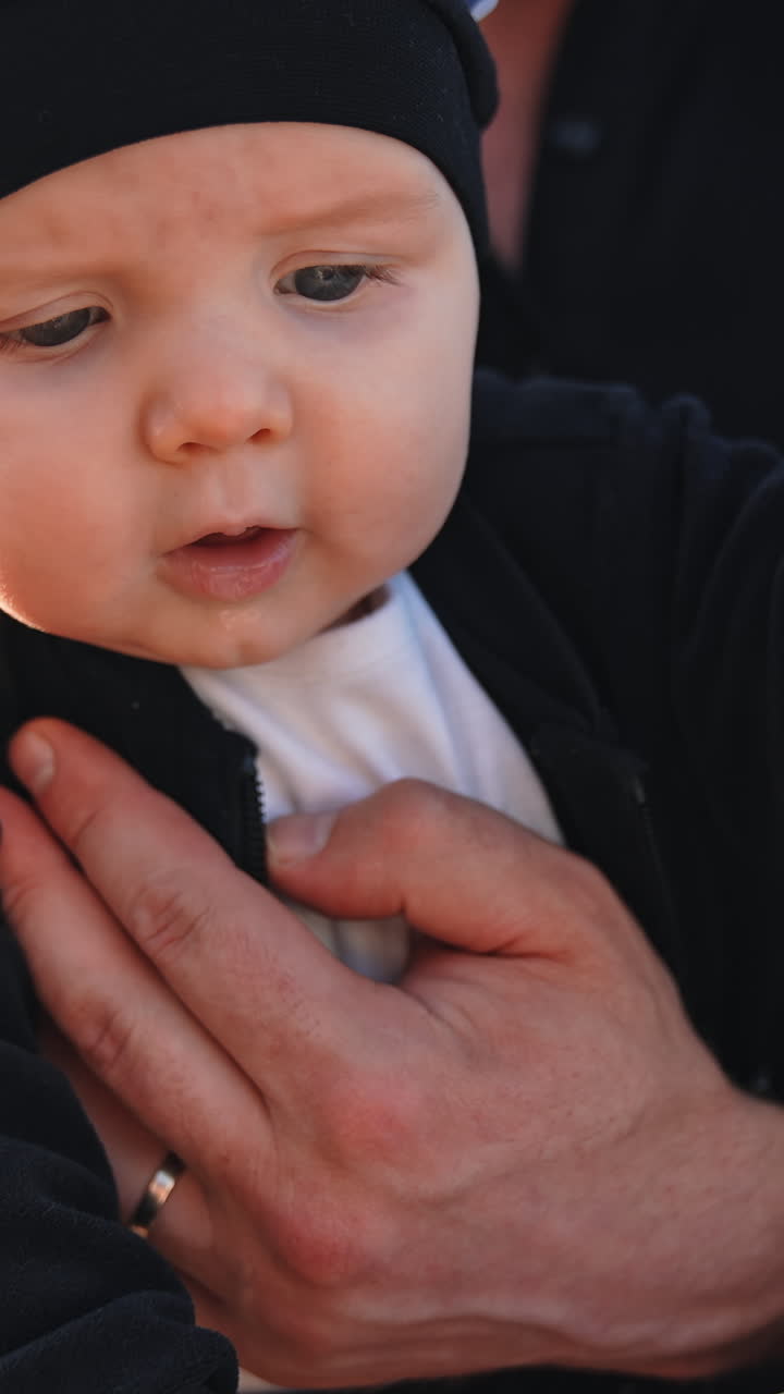 Cute little infant wearing black suit and cap. Baby in hands of parents is interested in the box in front of him. Close up. Vertical video