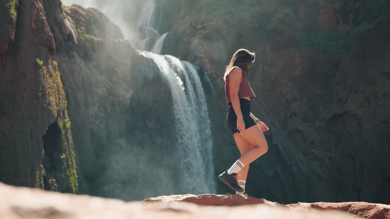 joven mujer caucásica caminando sobre rocas frente a las cascadas en ouzoud, marruecos