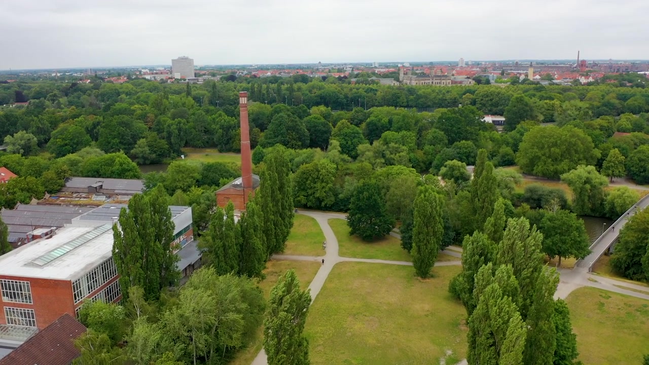 Uma vista aérea mostrando um exuberante parque verde em Hanôver, Alemanha, com uma antiga chaminé industrial imponente em meio às árvores e edifícios da cidade ao fundo.