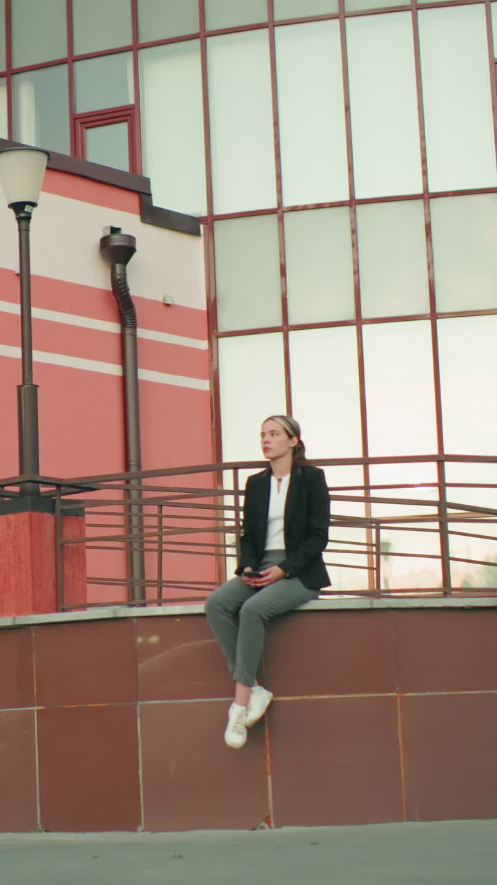 Calm lady in black blazer seated on fence with legs crossed at ankle holding phone, looking down in peaceful urban setting with car reflection on sleek tiles and modern glass building background