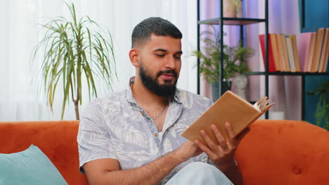indian man reading interesting book turning pages smiling enjoying literature taking a rest on sofa