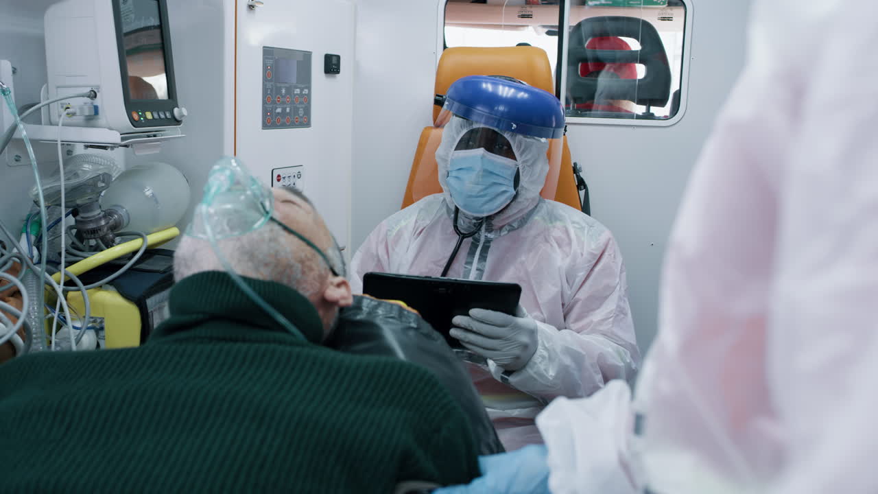 paramedic Using Tablet Near Coronavirus Patient Black Emergency Medicine Worker in Hazmat Suit Browsing Tablet Near Senior Patient with Oxygen Mask while Riding Ambulance during Pandemic