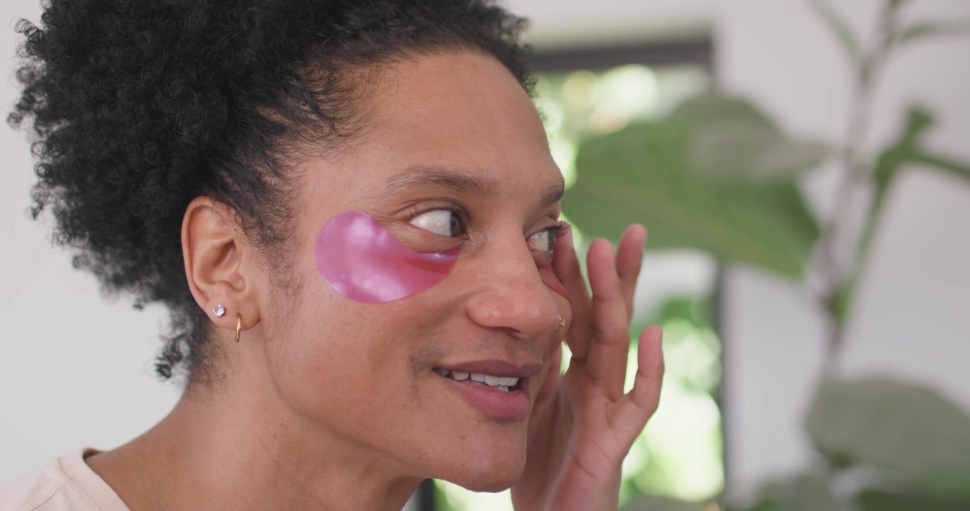 Male smiling, touching pink hydrogel eye patch, inspecting and smoothing placement in bright room