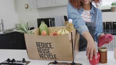 happy biracial woman unpacking groceries from box with recycle text in kitchen, slow motion