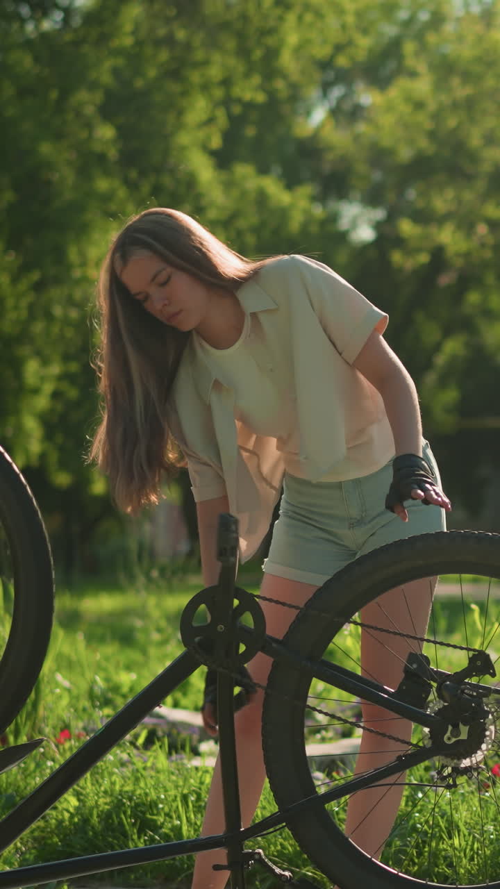 여성 자전거 타는 여성은 빛 공원 설정에서 활기찬 정원 에 거꾸로 자전거의 앞과 뒷 타이어를 조정합니다. 울창한 녹색 나무와 다채로운 꽃은 조용하고 흐릿한 배경을 제공합니다.