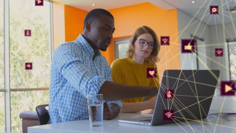 Animation-of-globes-of-digital-icons-over-diverse-man-and-woman-discussing-over-a-laptop-at-office