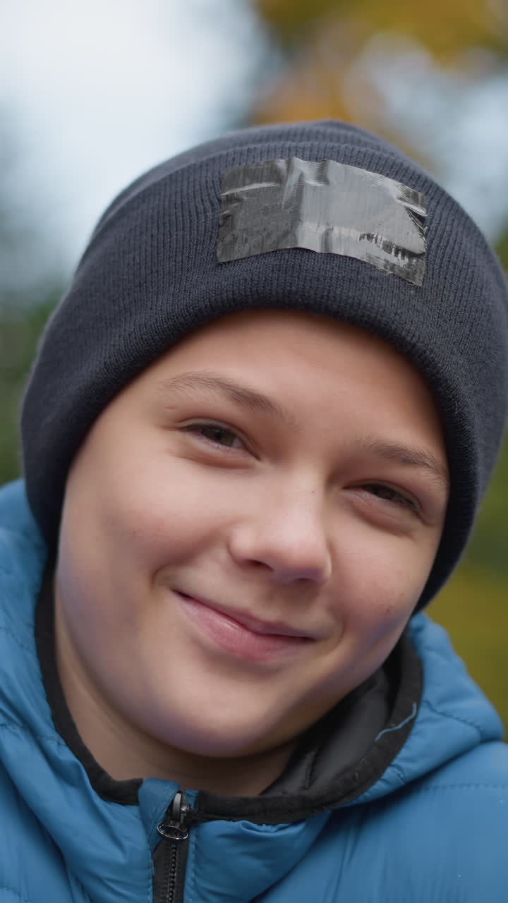 niño con gorra negra y chaqueta azul de pie al aire libre, sonriendo y disfrutando del momento, el fondo presenta vegetación borrosa y paisajes de otoño