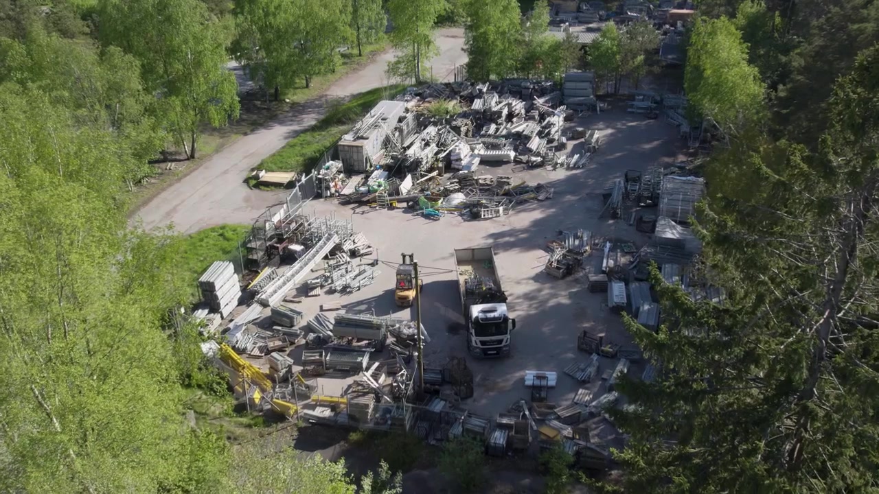 Aerial flyby of an industrial site where scaffolding frames and metal pipes are scattered across a storage yard.