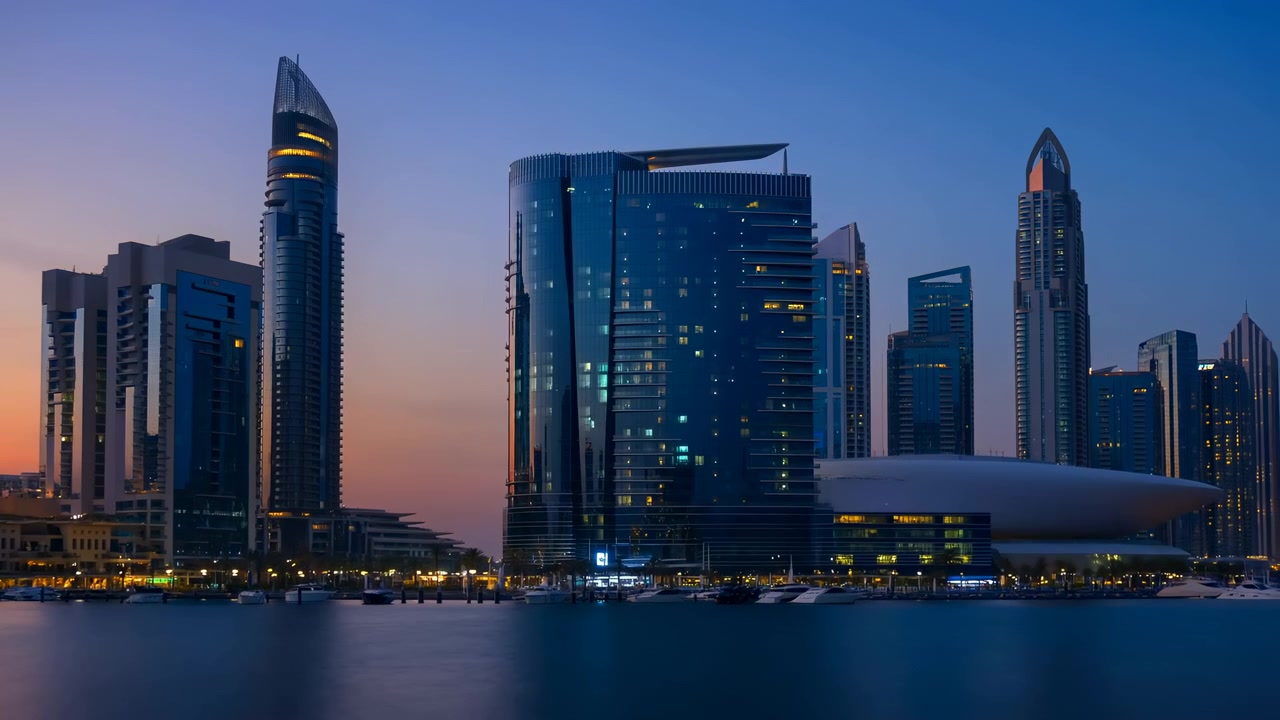 Panoramic waterfront city view showing lighting changing from sunset glow into twilight and night as modern towers, marina reflections, and glass facades brighten across urban shoreline