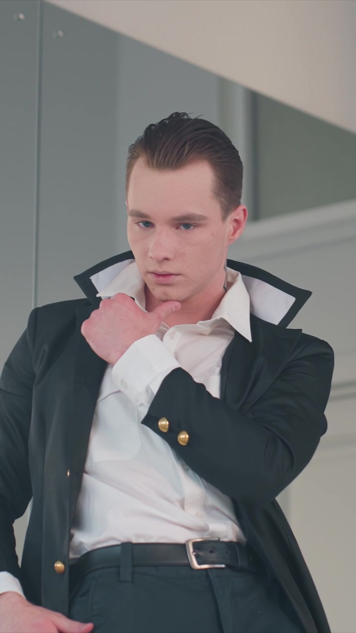 Giovane uomo in giacca nera con bottoni dorati e camicia bianca. I suoi capelli castani corti sono visibili mentre l'uomo sistema i dettagli dell'abbigliamento. Lo sfondo presenta pareti di colore chiaro.