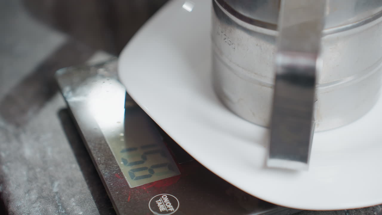 Close-up of stainless steel sifter placed on white plate atop digital kitchen scale displaying weight, kitchen setting with modern countertop, electronic scale with visible tare button
