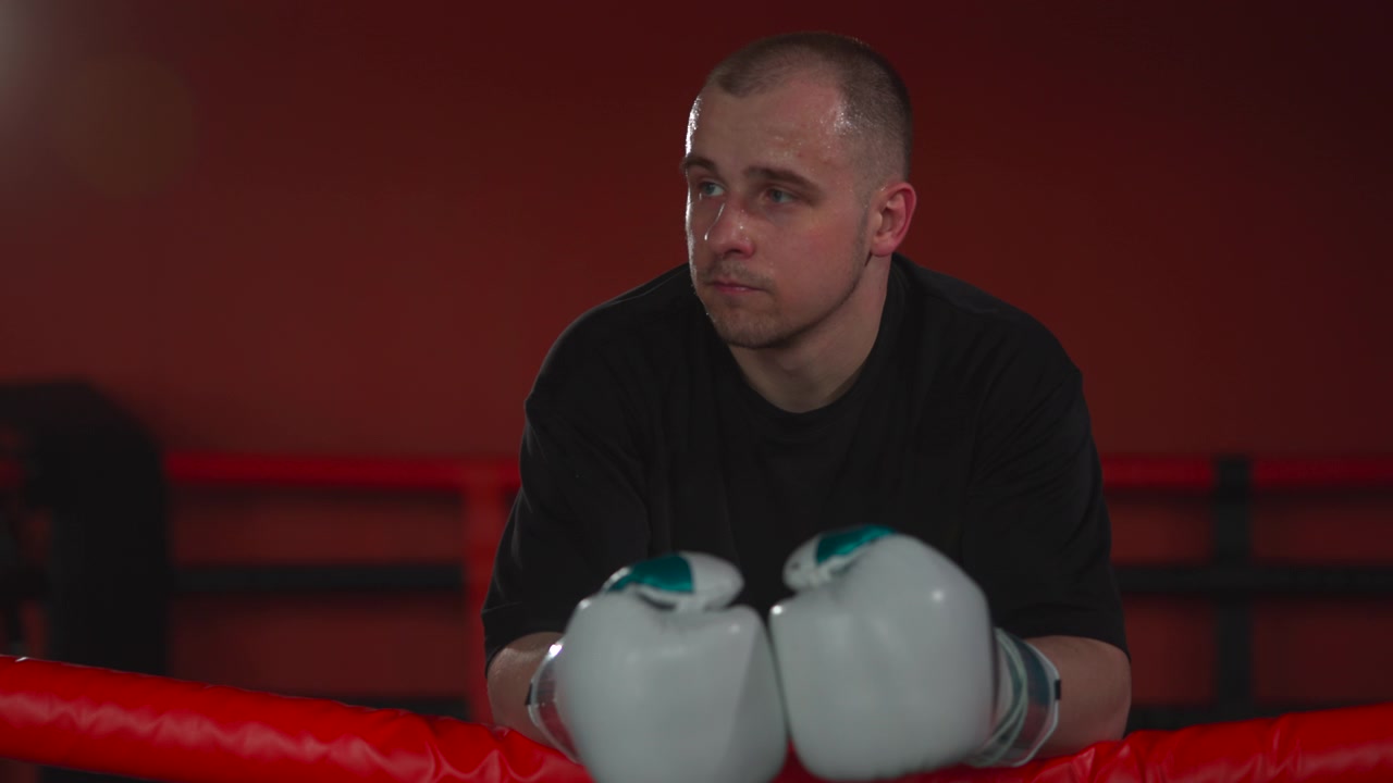 Focused young boxer rests on red ring ropes with white gloves raised in front, looking sideways inside dim indoor arena with red background, blurred railings, glossy highlights, and serious training