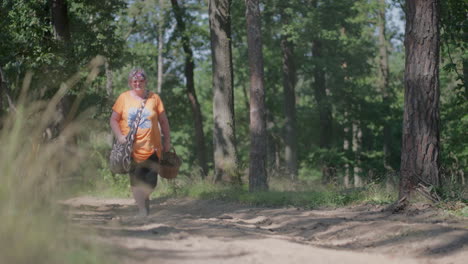 fat older woman with grey hair and orange t-shirt walking through the forest, holding wooden basket with a grass in the foreground, surrounded by trees during a day in slow motion