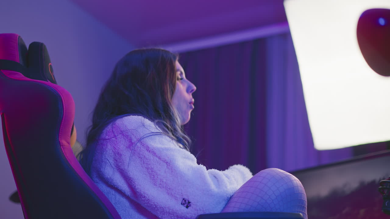 Side view of woman with long hair seated in ergonomic chair eating popcorn while watching screen, illuminated by bright studio lights with vibrant ambiance and microphone on desk