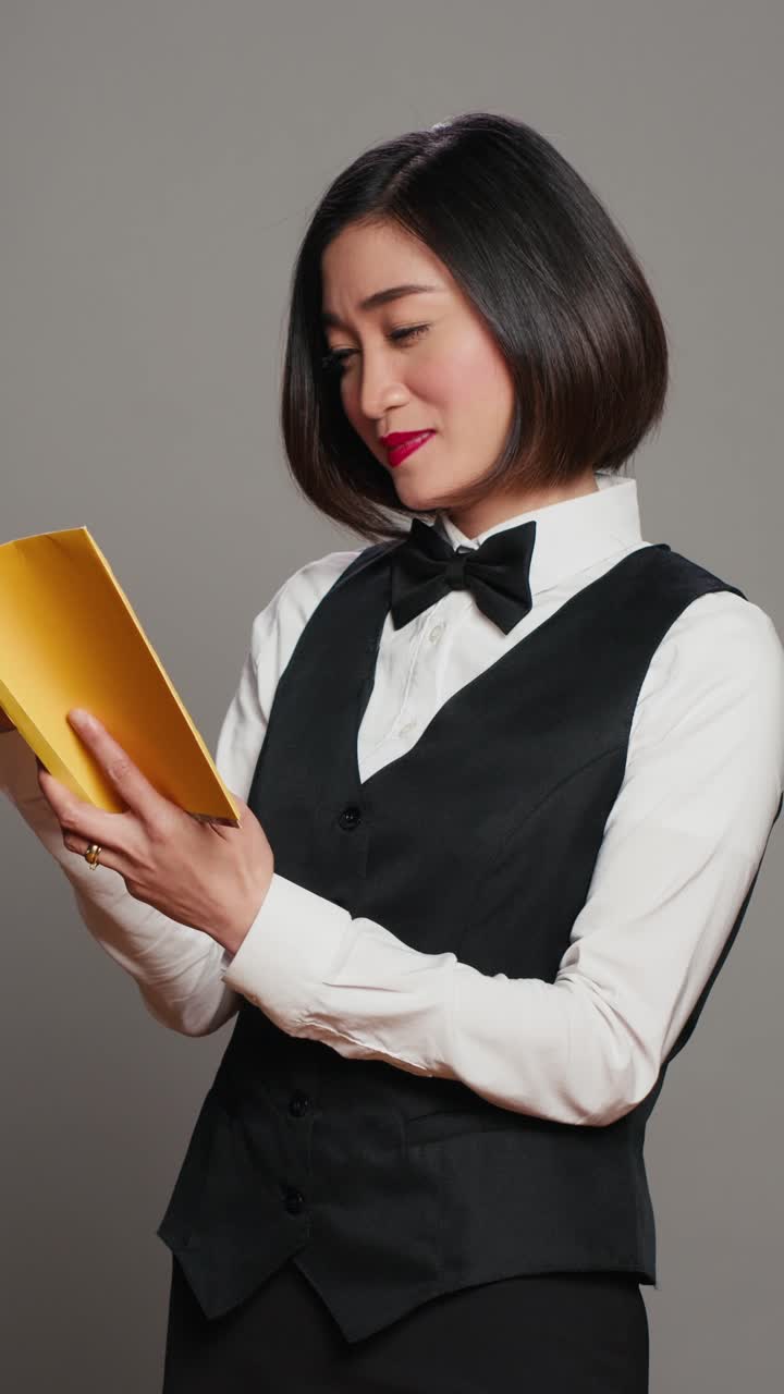 Video verticale di una receptionist asiatica che legge un libro di letteratura su sfondo grigio
