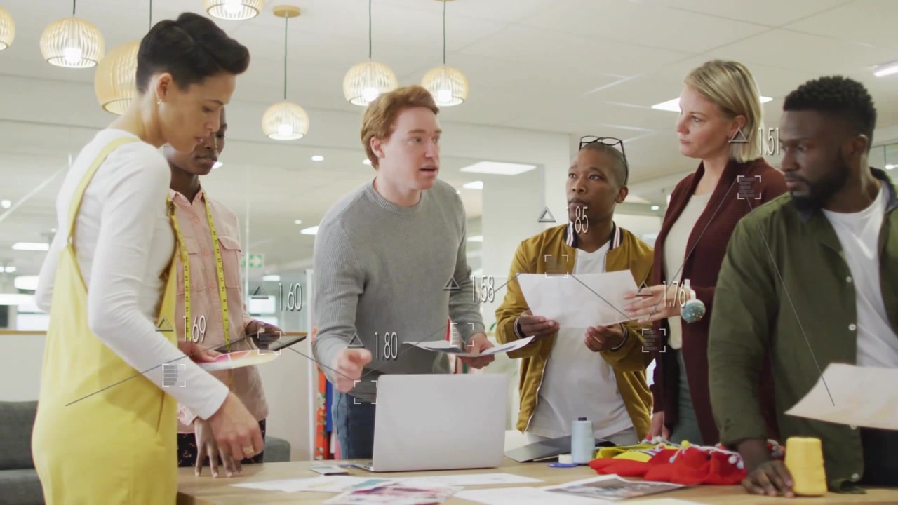 Leading biz-casual redhead man handing pages and guiding review at studio with laptop and swatches