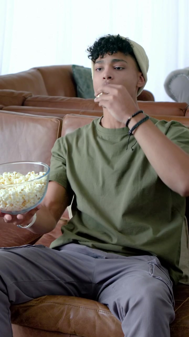 Video verticale: Uomo con berretto che prende popcorn da una ciotola di vetro su un divano in pelle per uno spuntino.