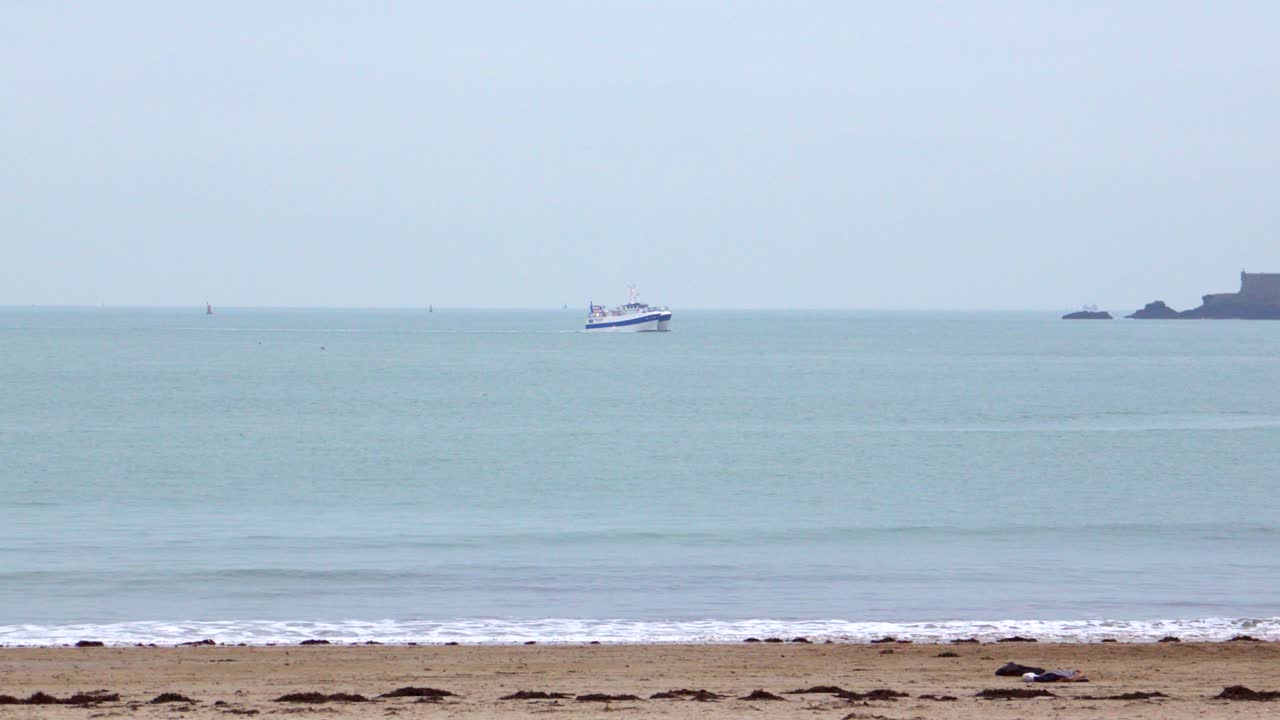 A slow-motion shot facing the open sea off Dinard. At the center of the frame, a small motorboat crosses the horizon under a grey winter sky, evoking calm and solitude.