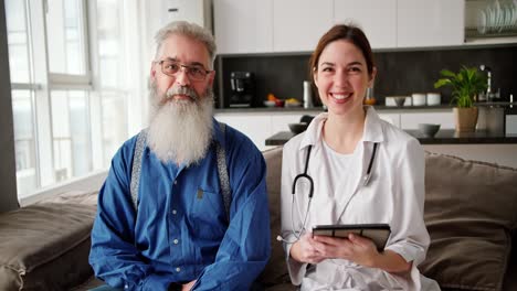 Portrait-of-an-elderly-man-with-a-lush-gray-beard-and-glasses-in-a-blue-shirt-who-sits-on-the-sofa-with-a-female-doctor-in-a-white-coat-who-holds-a-tablet-during-a-home-examination-in-a-modern-apartment