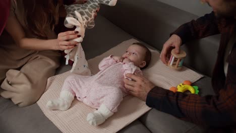 primer plano de una feliz joven morena junto con su joven marido jugando con su pequeña hija bebé en un mono rosa con la ayuda de una variedad de juguetes para niños en un sofá gris en un apartamento moderno
