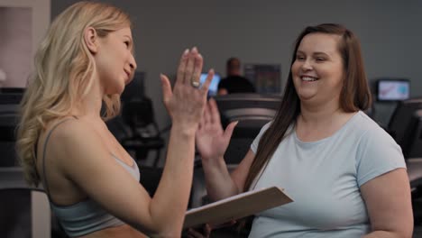 Eine-Kaukasische-Frau-Mit-Übergewicht-Macht-High-Five-Mit-Einer-Trainerin-Im-Fitnessstudio.