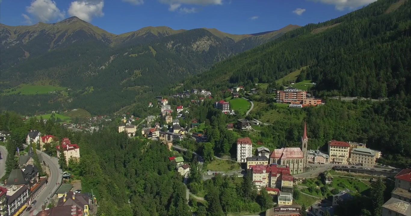 Lento volo laterale di un drone che rivela la chiesa di Bad Gastein, hotel storici, strade collinari tortuose, auto parcheggiate e case sparse sotto le verdi cime montuose austriache ricoperte di foreste