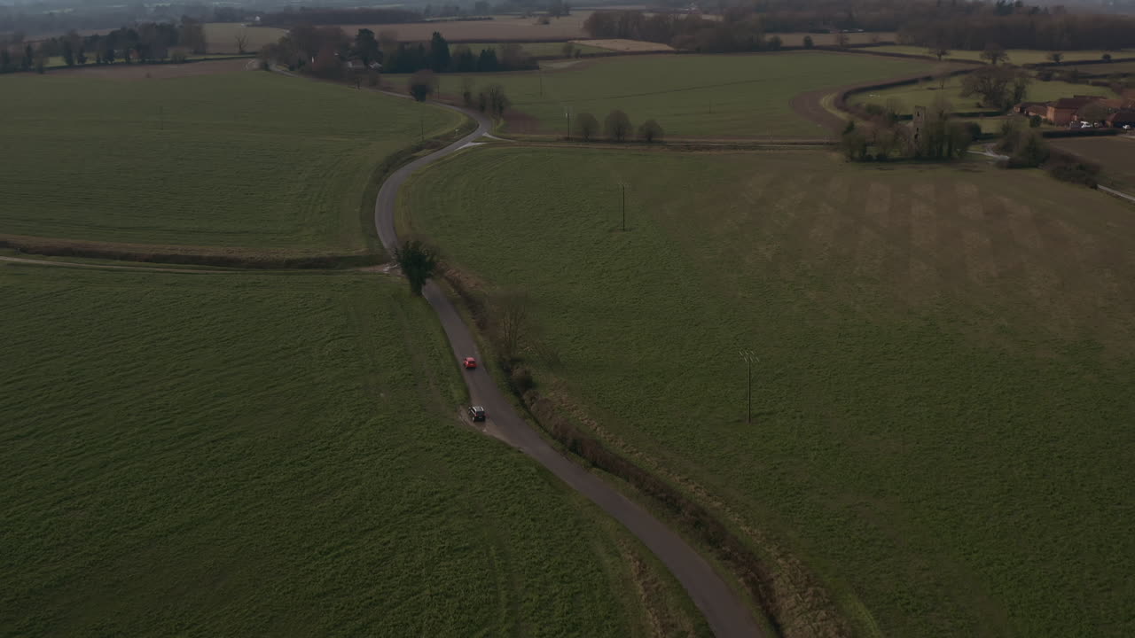 Aerial drone shot tracking a read card as it drives on a country lane surrounded by farms