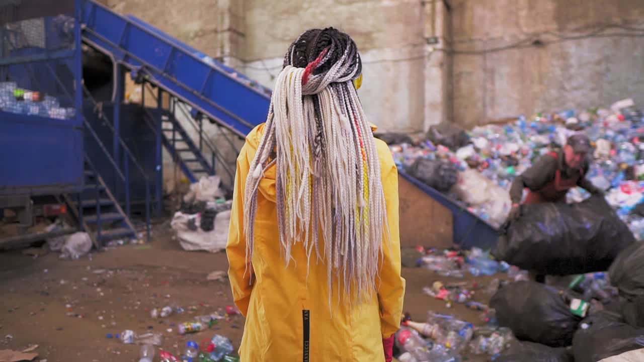 Backside footage of a woman with dreadlocks wearing yellow jacket, protective eyeglsses and mask standing against waste, garbage in recycling factory with worker and equipment on background